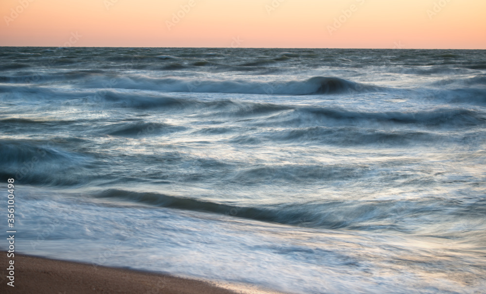 Fascinating sea waves splash along the sandy beach against a beautiful orange sunset on a warm summer evening. The concept of long summer days by the sea