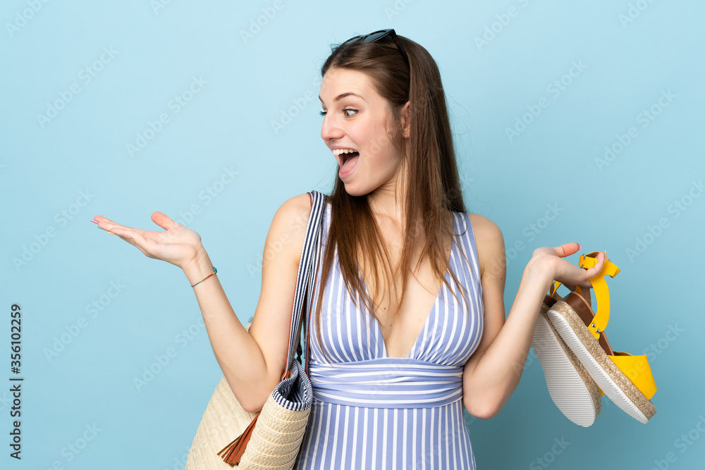 Young caucasian woman holding a beach bag an summer sandals isolated on blue background with surprise facial expression