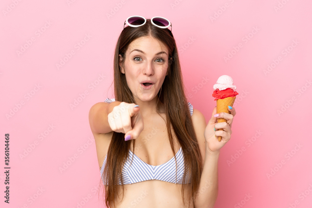 Young caucasian woman in swimsuit holding a cornet ice cream isolated on pink background surprised and pointing front Young caucasian woman in swimsuit holding a cornet ice cream isolated on pink background surprised and pointing front