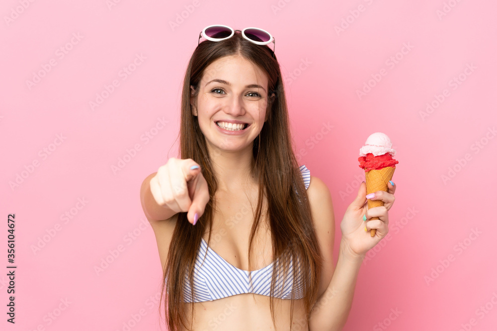 Young caucasian woman in swimsuit holding a cornet ice cream isolated on pink background points finger at you with a confident expression Young caucasian woman in swimsuit holding a cornet ice cream isolated on pink background points finger at you with a confident expression
