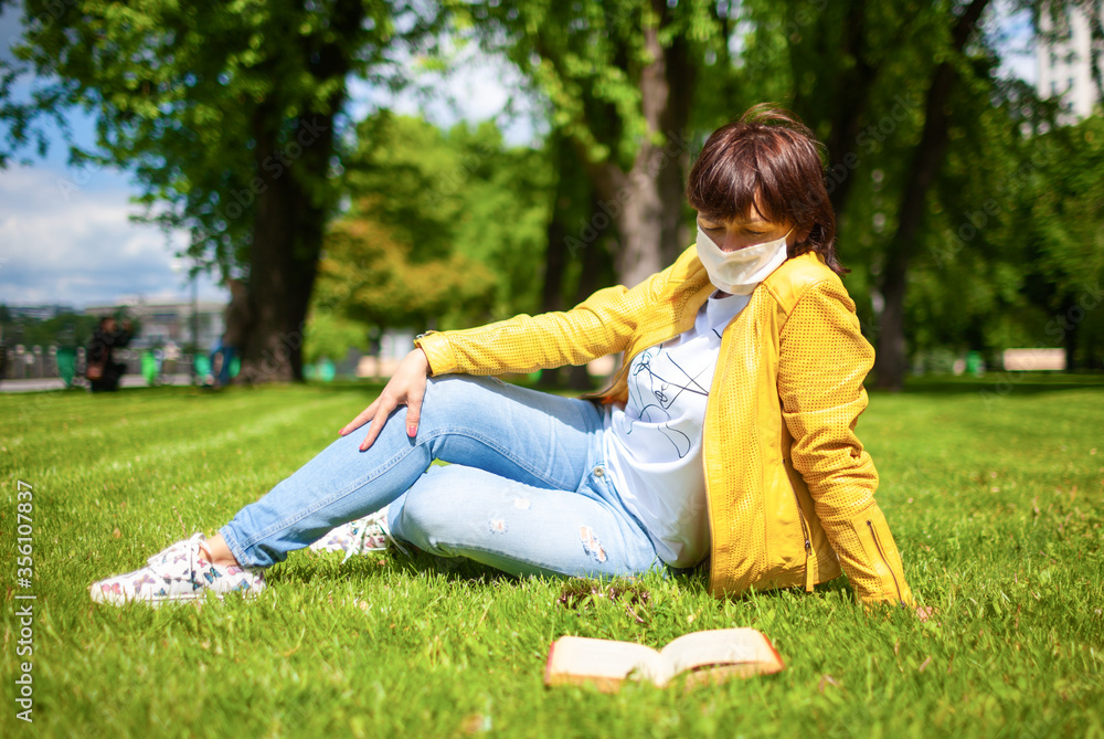 Side view of middle-aged caucasian woman putting white medical mask sitting on a green meadow in a sunny park next to a book. Coronovirus protection concept in the street