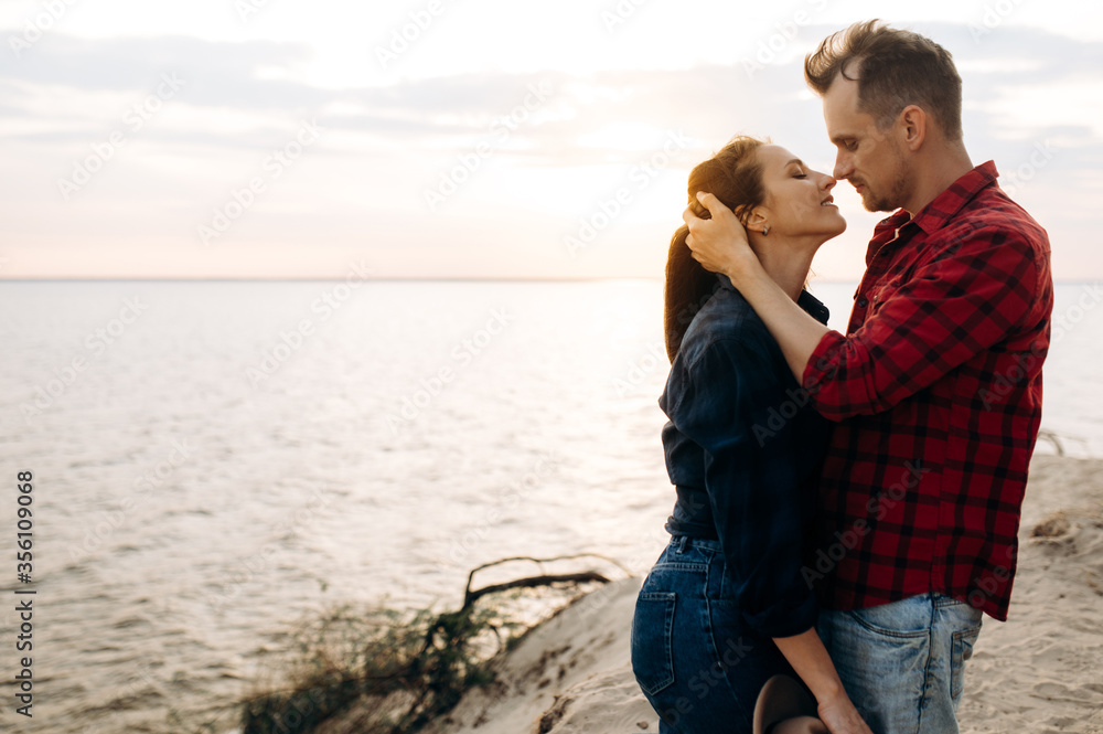 Young and happy couple in love dressed in stylish casual wear spend time together near the sea at sunset. The man gently hugs his beloved woman