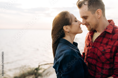 Young man and woman in love hug at sunset near the sea, they gently touch each other's noses and smiling. Romantic mood