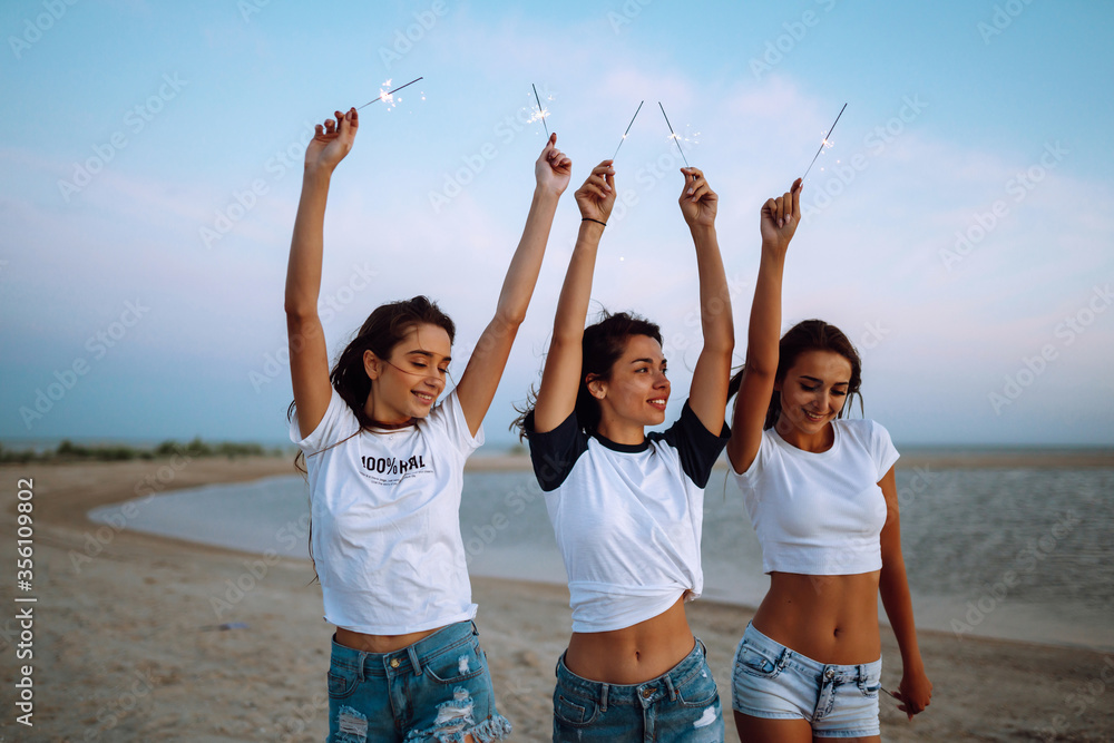 The sparklers in the hands of young girls on the beach. Three girls enjoying party on beach with sparklers. Summer holidays, vacation, relax and lifestyle concept. The sparklers in the hands of young girls on the beach. Three girls enjoying party on beach with sparklers. Summer holidays, vacation, relax and lifestyle concept.