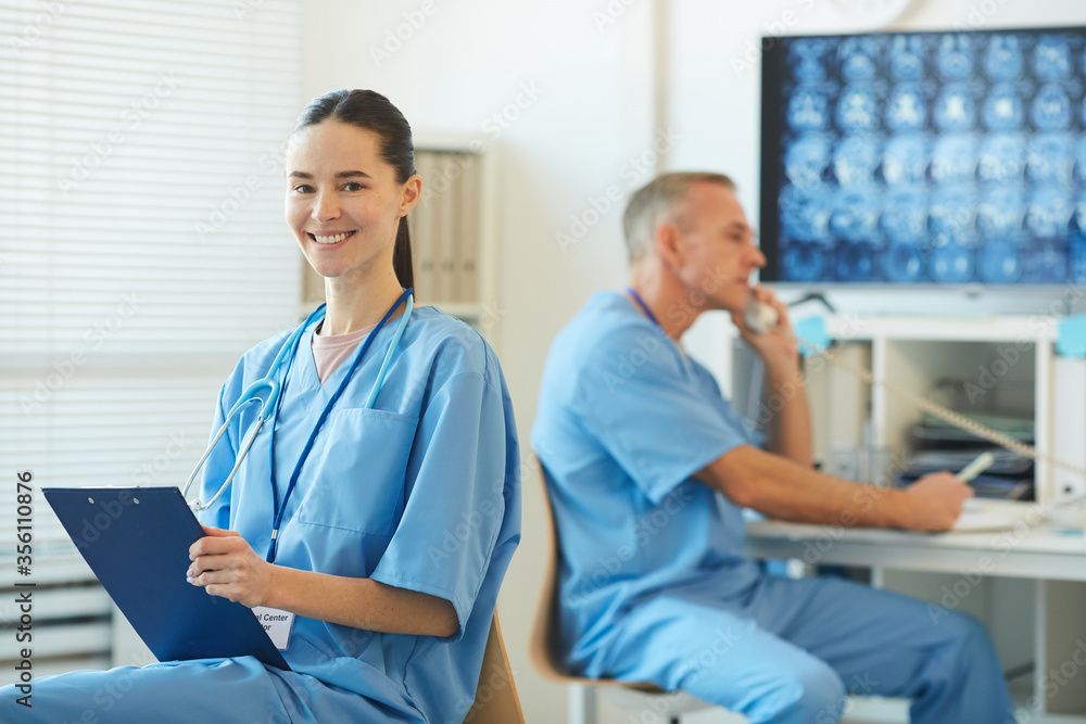 Obraz premium Waist up portrait of smiling female nurse holding clipboard and looking at camera while posing in clinic interior with mature doctor in background, copy space