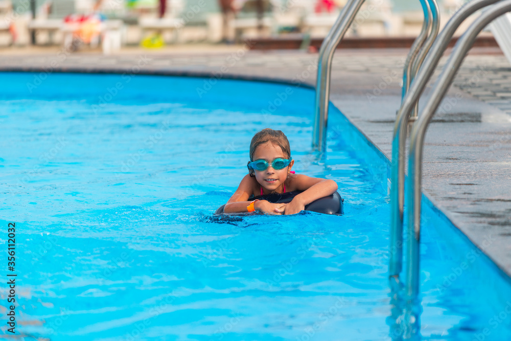 Charming little girl in swimming goggles and a lifebuoy swims in the clear blue water of the pool during summer vacation. Concept of children's holidays