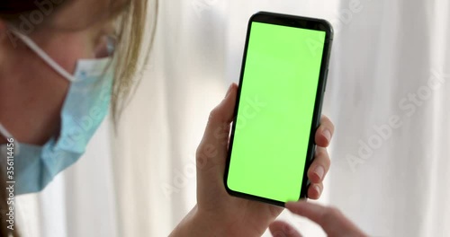 Side view of crop female in medical face mask focusing on green screen and interacting with smartphone against blurred white surface