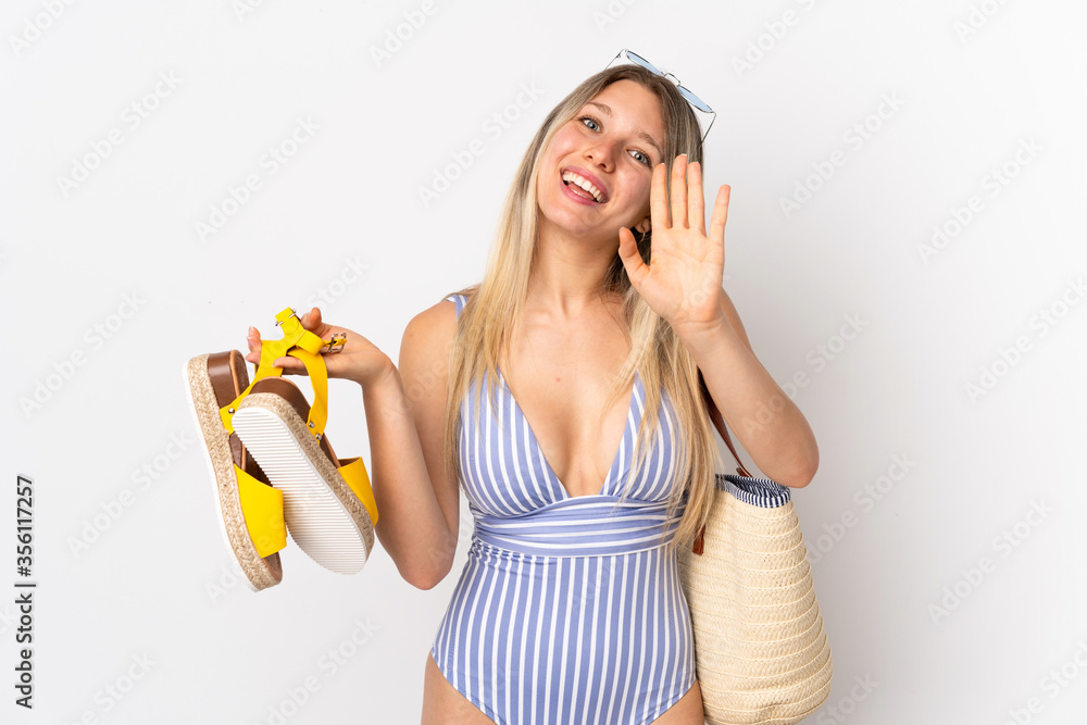 Young blonde woman holding summer sandals isolated on white background saluting with hand with happy expression Young blonde woman holding summer sandals isolated on white background saluting with hand with happy expression