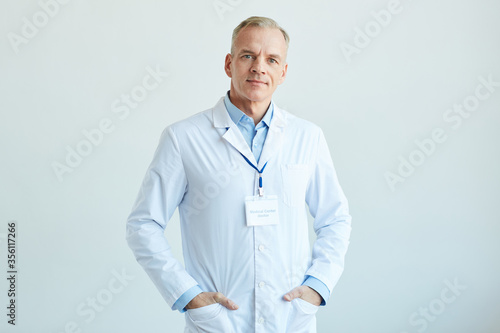 Waist up portrait of confident mature doctor wearing lab coat and looking at camera while standing against white wall in clinic, copy space photo