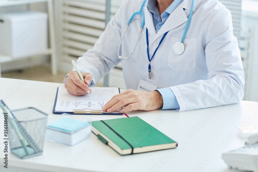Fototapeta premium Portrait of unrecognizable doctor writing on clipboard and wearing lab coat while sitting at desk in office of modern clinic, copy space