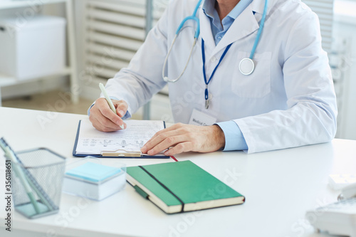 Portrait of unrecognizable doctor writing on clipboard and wearing lab coat while sitting at desk in office of modern clinic  copy space