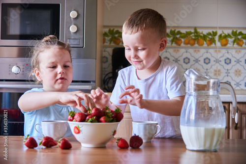 Two beautiful children in the kitchen eating strawberries and drinking milk.Children have fun. 