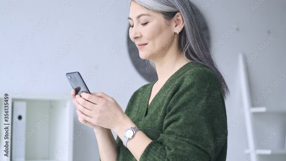 A good-looking mature businesswoman with long gray hair is using her smartphone while working in the office A good-looking mature businesswoman with long gray hair is using her smartphone while working in the office