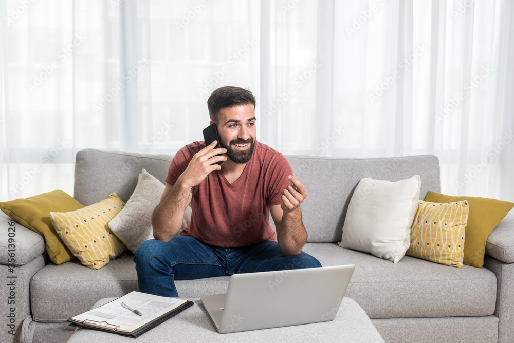 Young bearded freelancer man sitting on the sofa talking on the phone and working on his laptop with smile on his face