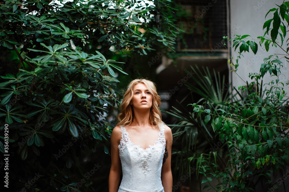 Portrait of an attractive blonde bride standing in a wedding dress on a background of greenery. The bride in the botanical green garden. Beautiful young woman with curly blond hair. Marriage. Portrait of an attractive blonde bride standing in a wedding dress on a background of greenery. The bride in the botanical green garden. Beautiful young woman with curly blond hair. Marriage.