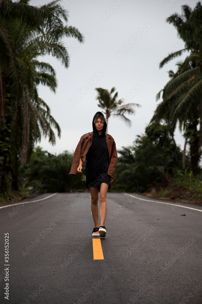 Girl in a brown leather jacket is on a paved road with a yellow stripe amongst the palm trees
