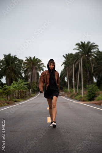 Girl in a brown leather jacket is on a paved road with a yellow stripe amongst the palm trees