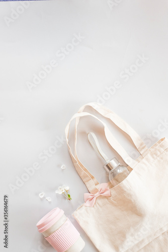 Zero waste shopping concept - cotton bag with pink bow, reusable cup for tea coffe with spring flowers, herbs and leaves on a light background.