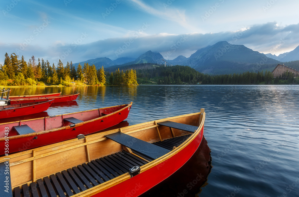 Beautiful sunny landscape. View on mountain lake with crystal clear azure water in High Tatras. Slovakia. Red Boats on the water glowing in sunlight at sunset. Awesome Autumn landscape. Strbsce pleso
