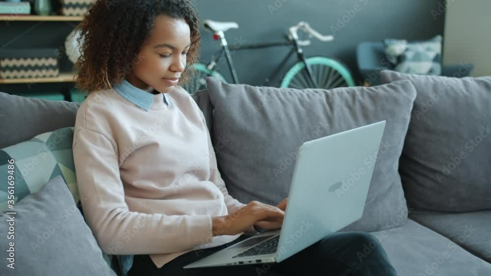 African American girl is using laptop in apartment smiling typing enjoying freelance work sitting on couch indoors . Electronic devices and freelancers concept. African American girl is using laptop in apartment smiling typing enjoying freelance work sitting on couch indoors . Electronic devices and freelancers concept.