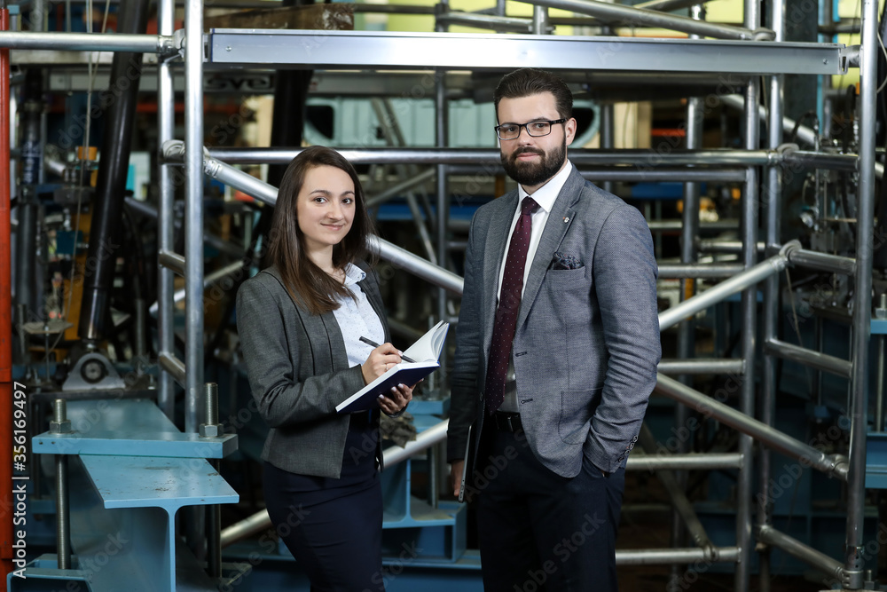 Portrait of a solid businessman with his secretary holding blue notebook, talking about factory financial report in a airplane manufactory. Factory managers controlling work process in a factory Portrait of a solid businessman with his secretary holding blue notebook, talking about factory financial report in a airplane manufactory. Factory managers controlling work process in a factory