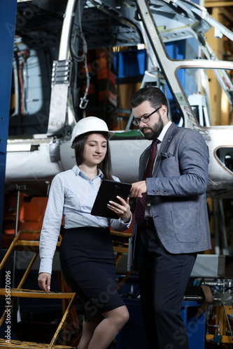 Portrait of a solid businessman with his secretary holding laptop and tablet, talking about factory financial report in a aircraft manufactory.