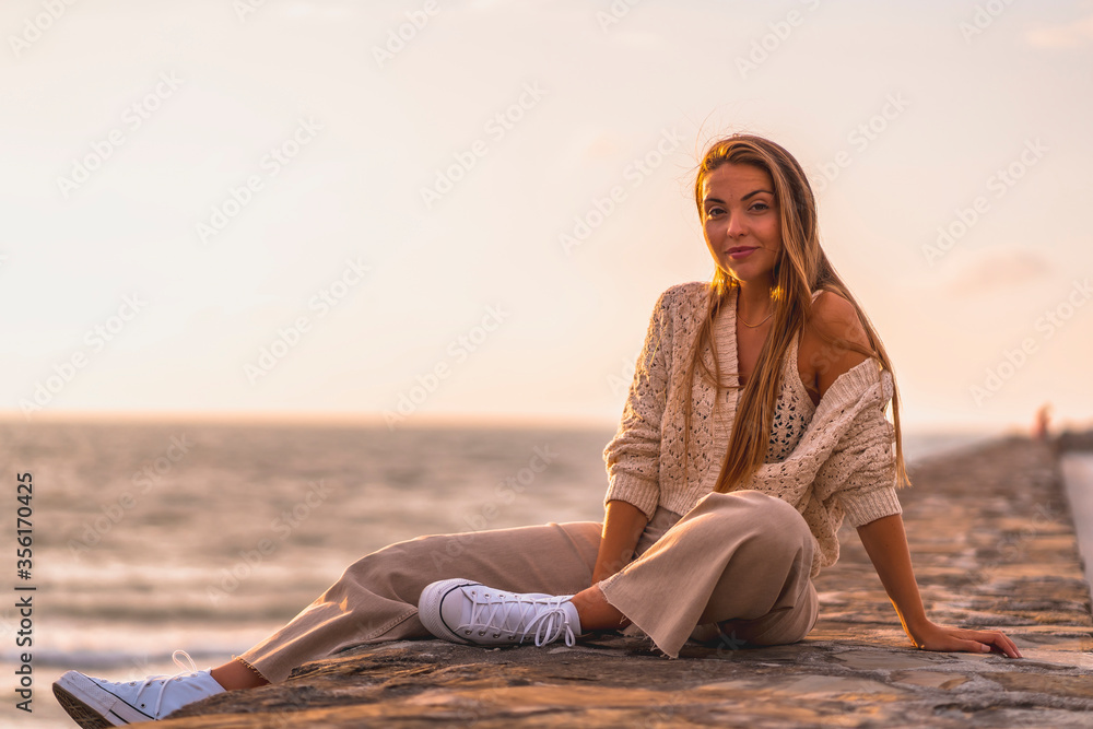 Summer lifestyle, a young blonde Caucasian woman sitting by the sea in a white crop top and corduroy pants. On a summer afternoon