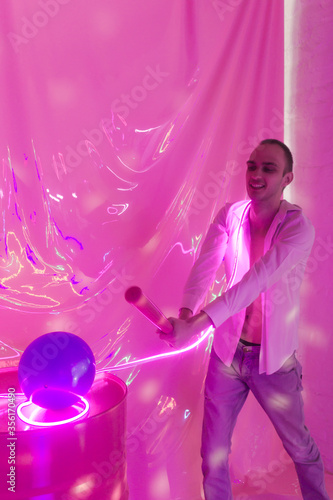 Portrait of the Young Handsome emotional man in an unbuttoned white shirt with bat breaks the ball. Neon Room. Old 90's retro style. Reflect of neon words near male. Selective focus.