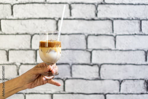 Coffee Macchiato and milk layer with ice in champagne glass was held by Asian grandmother hand in front of white brick wall wallpaper.