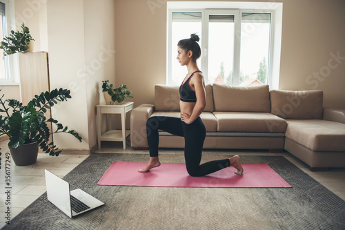 Fototapeta Naklejka Na Ścianę i Meble -  Young brunette woman wearing sportswear is doing yoga on a pink carpet using a laptop