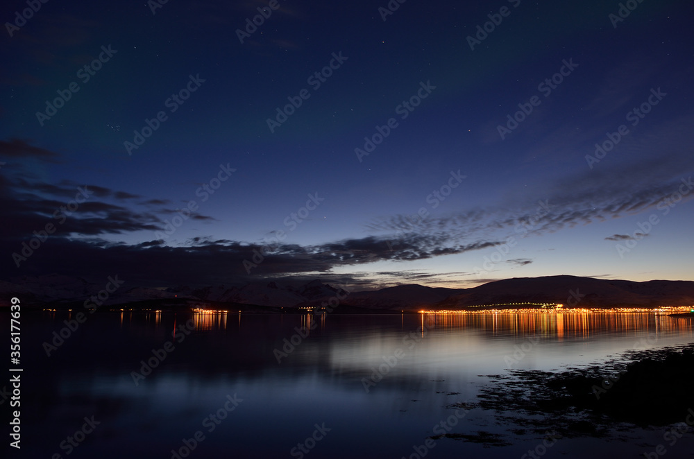 calm reflective fjord surface at night time with city lights in the background