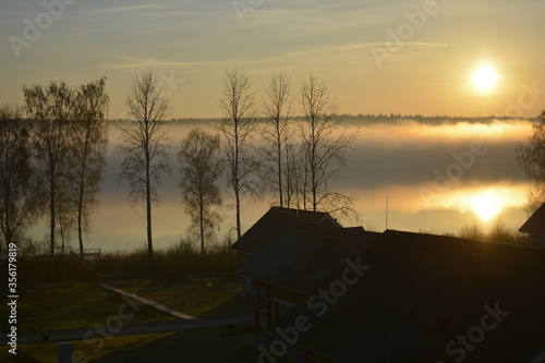 Leningradskaya oblast Russia, May 05 2016. Sunrise over the lake in the fog. Smoke over water and silhouettes of small houses and trees photo