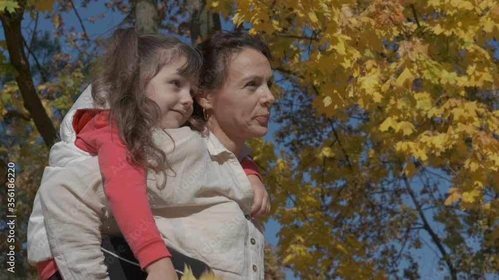 Mother hold child in forest. A cute little girl sits on her mother's back in an autumn park. Mother hold child in forest. A cute little girl sits on her mother's back in an autumn park.