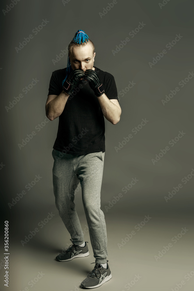 A man with an unusual hairstyle of mohawk from blue braids in boxing gloves poses and is ready for battle on a dark background. A man with an unusual hairstyle of mohawk from blue braids in boxing gloves poses and is ready for battle on a dark background.