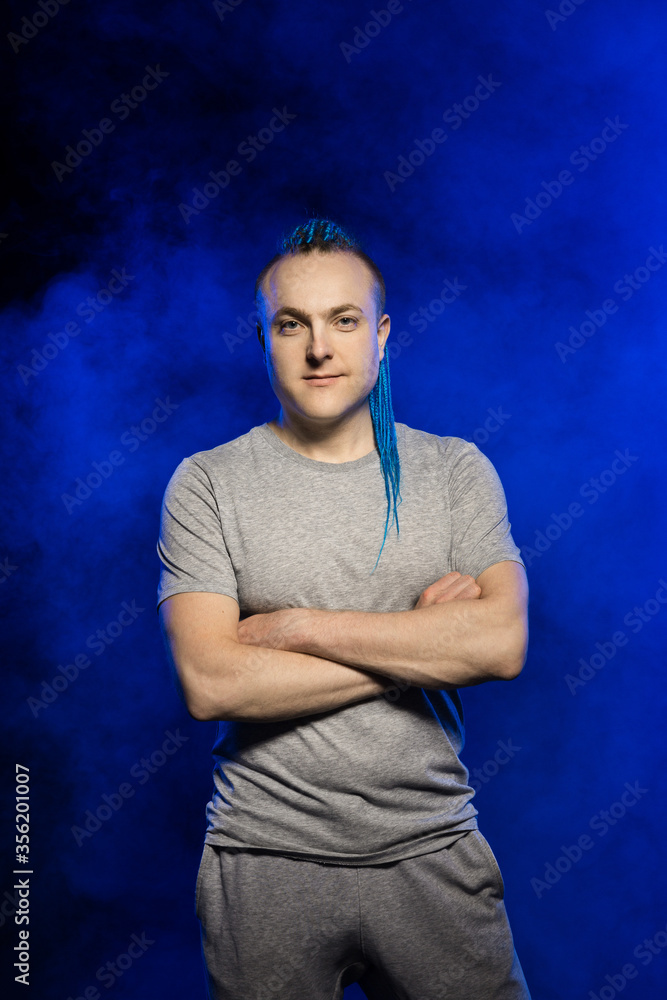 A man with a braided mohawk haircut calmly posing arms crossed against a background of blue smoke.