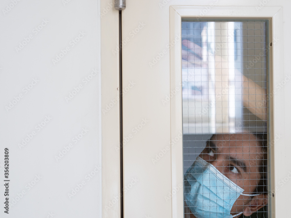 Lonely man in medical mask looking through the window. Isolation at home for self quarantine. Concept home quarantine, prevention COVID-19. Coronavirus outbreak situation Lonely man in medical mask looking through the window. Isolation at home for self quarantine. Concept home quarantine, prevention COVID-19. Coronavirus outbreak situation