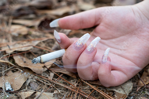 A female hand holding a burning cigarette with a smoke trail on the ground  surrounded by fallen brown leaves  CIGARETTE 