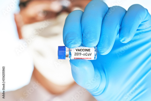 Hand in blue medical gloves holding small vial with label (own design, not real product) 2019-nCoV vaccine, blurred face in cotton mask and goggles background. Coronavirus covid 19 cure concept