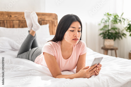 Asian Girl Playing Game On Smartphone Lying On Bed Indoors