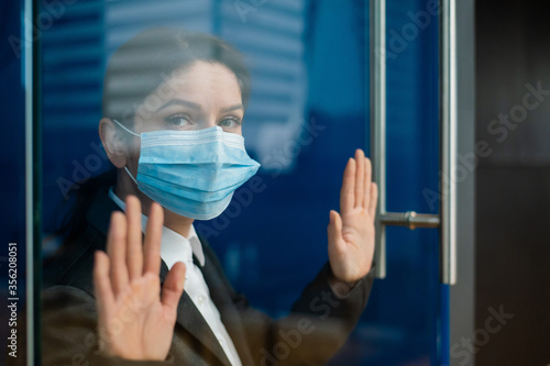 A sad quarantined woman in a business suit leaned against the window. Caucasian girl in a mask on isolation put her hands on a glass door. Maintaining social distance to prevent coronavirus.