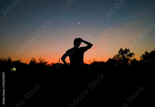 Silhouette of a boy scratching his head in confusion and thinking in the evening with crescent moon in the sky after sunset as background. Inspirational. De clutter mind concept. Depression.