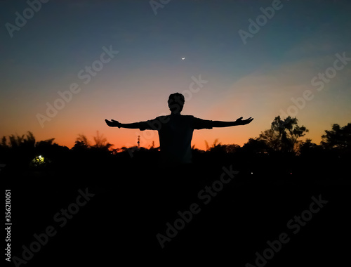 Silhouette of a man expressing freedom and love with stretched arms or hands in praise in the evening with crescent moon in the colorful sky after sunset as background. Inspirational. Freedom concept.
