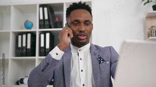 Likable purposeful experiencedbusy dark-skinned businessman talking on phone with business partner while working on computer in contemporary office photo