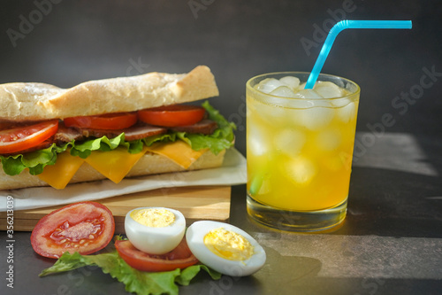 big tasty sandwich with tomato lettuce with soap and ham on the board. next to it is juice with ice in a glass.