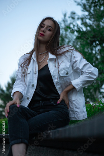 a teenage brunette girl in a white jacket is sitting on a bench in the Park. Bright red lipstick adds to the attractiveness and charm.