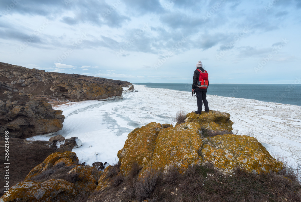 Obraz premium Tourist girl with a red backpack standing on a rock and looking at the frozen sea. Winter landscape. Solitude in the wilderness