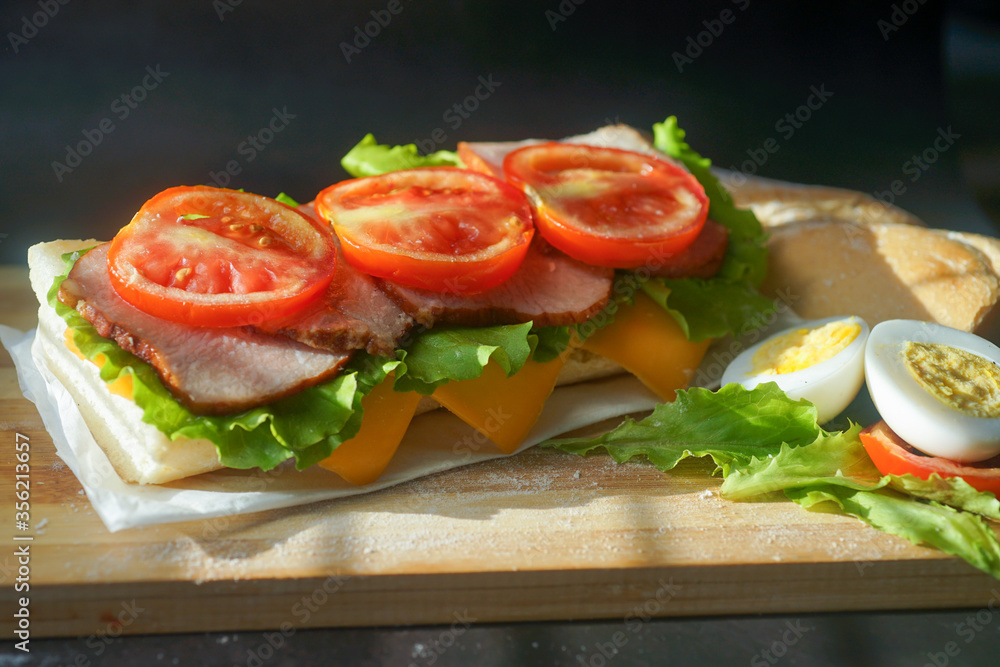 An appetizing sandwich with ham, cheese, tomatoes and lettuce lies on a board with egg and lemon on black background An appetizing sandwich with ham, cheese, tomatoes and lettuce lies on a board with egg and lemon on black background