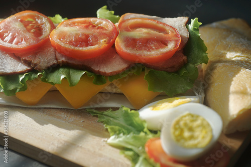 An appetizing sandwich with ham, cheese, tomatoes and lettuce lies on a board with egg and lemon on black background
