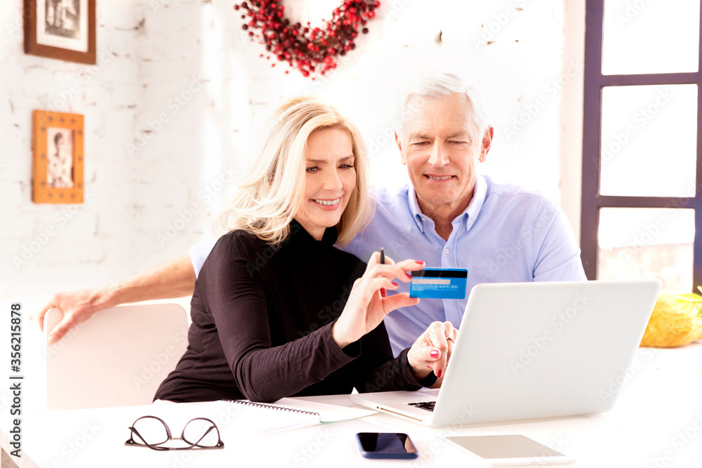 Happy elderly couple using bank card and notebook while shopping online at home Happy elderly couple using bank card and notebook while shopping online at home