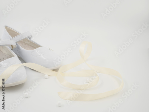 Pair of white baby lacquered girls shoes with beige ribbon and white balls at white background with place for text